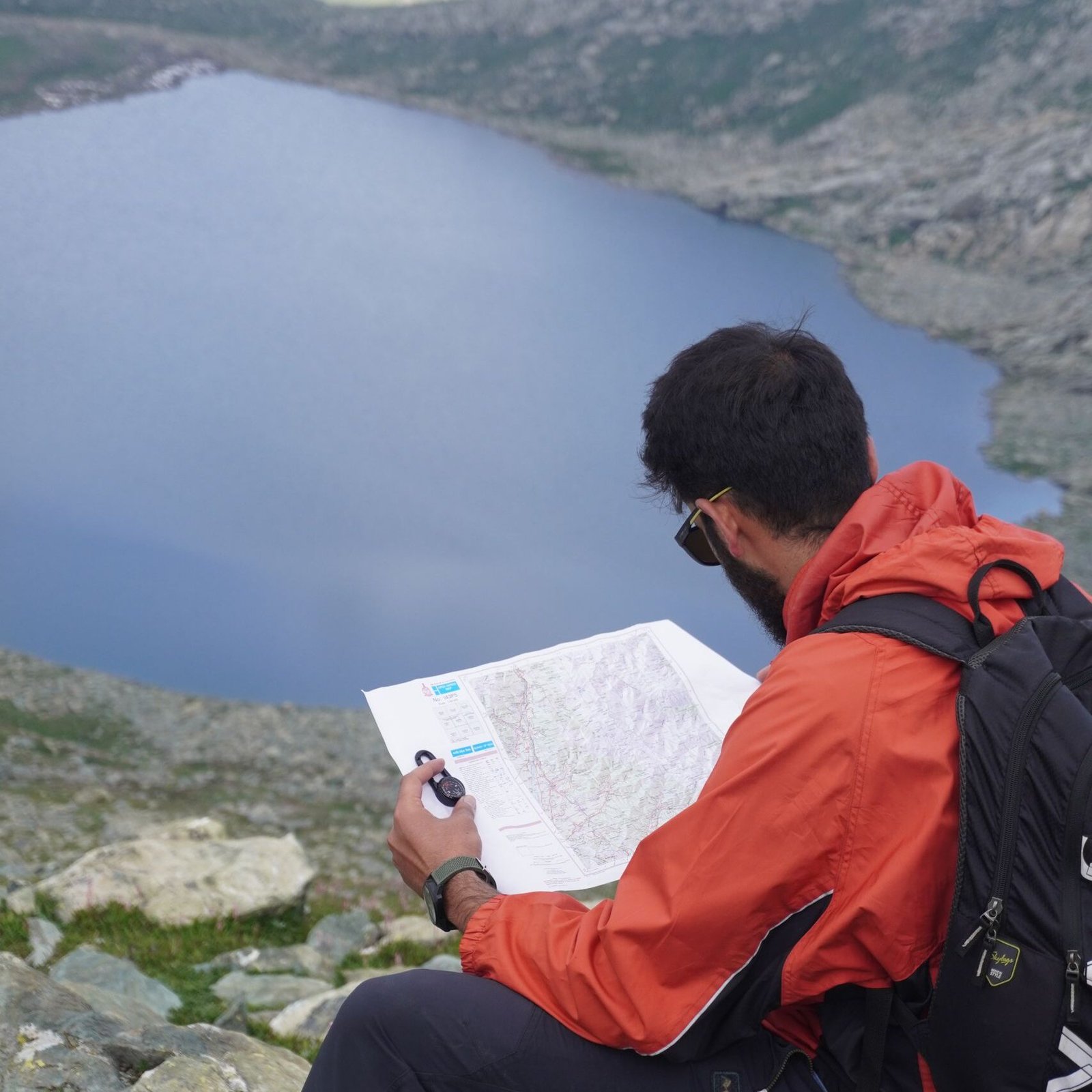 A hiker in orange jacket studies a map overlooking a mountain lake.